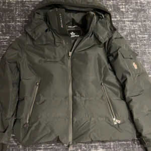Svart vinterjacka från Moncler - Snygg svart dunjacka från Moncler med avtagbar huva och dragkedja framtill. Jackan har två snedställda fickor med dragkedja och en liten ficka på ärmen. Perfekt för kalla dagar.