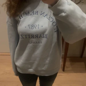 Ljus blå sweatshirt med tryck - Säljer en ljus blå sweatshirt med blått tryck på framsidan. Tröjan har långa ärmar och en avslappnad passform. 