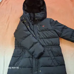 Säljer en svart dunjacka från Levi's med huva och dragkedja. Jackan har en klassisk design med quiltade paneler och en liten röd Levi's-logga på bröstet. Perfekt för kyliga dagar.