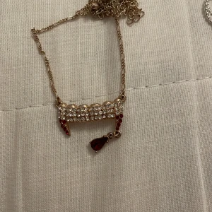 Guldhalsband med röda och klara stenar - Elegant guldhalsband med en rad av klara stenar och hängande röda droppformade stenar. Perfekt för att ge en touch av glamour till din outfit.