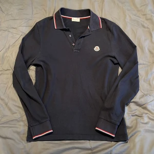 Marinblå långärmad pikétröja från Moncler - Säljer en stilren mörkblå långärmad pikétröja från Moncler. Den har en klassisk krage med knappar och snygga detaljer i rött och vitt på krage och ärmslut. Perfekt för både vardag och fest!