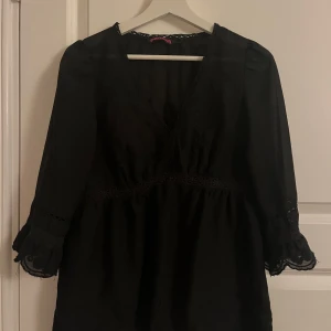 Svart blus med spetsdetaljer - Elegant svart blus med v-ringning och spetsdetaljer vid ärmslut och midja. Blusen har trekvartsärmar och en lös passform, perfekt för en stilren look.