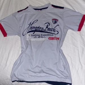 Vit baby tee med tryck från Hampton Beach - Snygg vit t-shirt från Hampton Beach med blått och rött tryck framtill. Den är ganska kort i magen och går under naveln precis, ungefär i storlek s/sx runt nacken o ärmarna. 🫶‼️