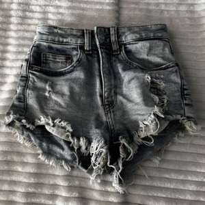 Grå jeansshorts med fransar - Säljer ett par coola grå jeansshorts med hög midja och slitna fransdetaljer. Perfekta för en avslappnad sommarlook. De har klassiska fem fickor och en knappgylf.⚠️ Ni betalar för frakt⚠️