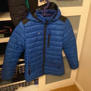 Blå jacka från Craft - Snygg blå jacka från Craft med svarta axelpartier och huva. Jackan har dragkedja framtill och praktiska fickor med dragkedjor. Perfekt för kyligare dagar.