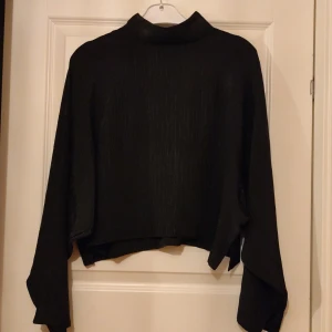 Svart långärmad blus med knappar - Snygg svart topp med hög krage och knappar i nacken. Den har ett subtilt randigt mönster och långa ärmar. Perfekt för en stilren look.