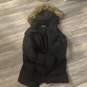 Svart vinterjacka med pälsdetalj från Indigo - Säljer en svart vinterjacka från Indigo med pälskantad huva. Jackan har en pufferdesign och är perfekt för kalla dagar. Den är knappt använd, storleken är M