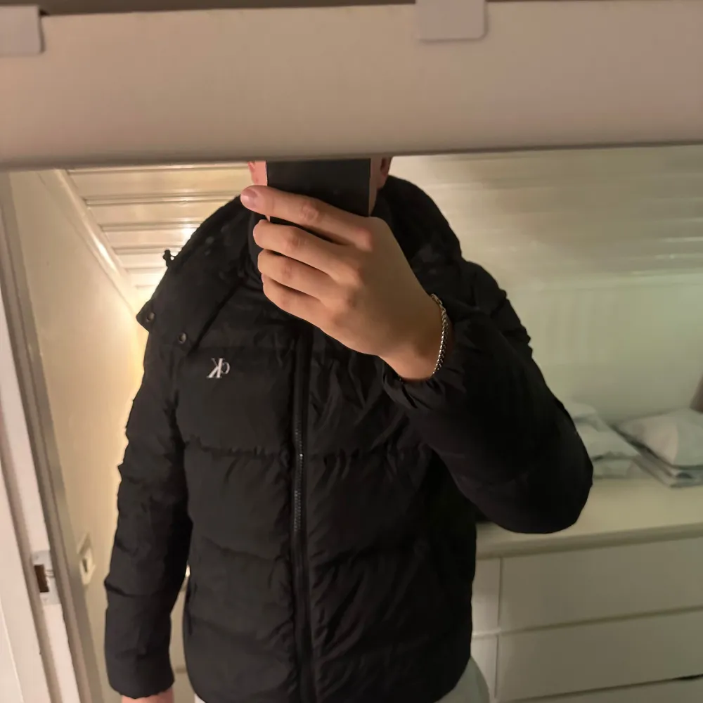 Säljer en snygg svart dunjacka från Calvin Klein i mycket bra skick. Jackan har en dragkedja framtill och en mysig huva. Perfekt för kalla vinterdagar och har en stilren design med CK-logga på ärmen. Passar både till vardags och lite finare tillfällen. I strl L. Takit.