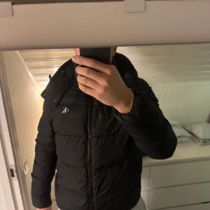 Svart dunjacka från Calvin Klein - Säljer en snygg svart dunjacka från Calvin Klein i mycket bra skick. Jackan har en dragkedja framtill och en mysig huva. Perfekt för kalla vinterdagar och har en stilren design med CK-logga på ärmen. Passar både till vardags och lite finare tillfällen. I strl L