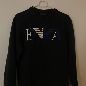 Svart tröja från Emporio Armani - Säljer en stilren svart tröja från Emporio Armani med deras ikoniska logga i vitt och blått på bröstet. Tröjan har långa ärmar och är perfekt för både vardag och fest. Passar bra till jeans eller chinos för en avslappnad men snygg look.