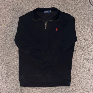 Svart tröja från Polo Ralph Lauren - Säljer en stilren svart tröja från Polo Ralph Lauren med en halv dragkedja och det klassiska röda logotypbroderiet på bröstet. Perfekt för både vardag och lite finare tillfällen. Tröjan har långa ärmar och är i mycket bra skick. Passar perfekt till höst och vinter!