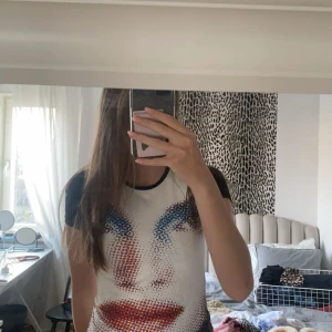 Vit och svart t-shirt med ansiktsmönster - Säljer en cool t-shirt med ett unikt ansiktsmönster i rött och blått på framsidan. T-shirten är vit med svarta ärmar och har en normal passform. Perfekt för en avslappnad stil eller en utekväll. Skicka ett meddelande vid intresse!