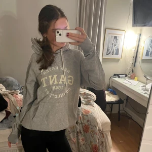 Grå hoodie från GANT - Säljer en grå hoodie från GANT med tryck på framsidan. Den har en klassisk känguruficka och justerbar huva med snören. Perfekt för en avslappnad stil.