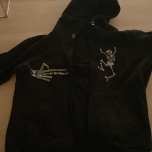 Svart hoodie med skelettmotiv - Rhinestone hoodie i svart med dragkedja och huva. Den har ett unikt skelettmotiv i glittrande stenar på bröstet och ett mönster på ärmen. I storlek S. Passar perfekt för mig som är 180 lång.