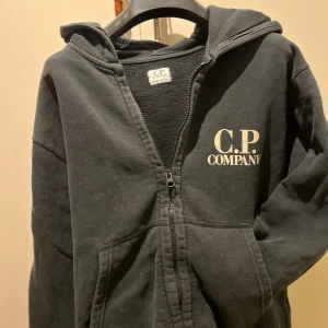 Svart C.P. Company Hoodie - Säljer en svart hoodie från C.P. Company med dragkedja och unika glasögondetaljer på huvan. Perfekt för en cool och avslappnad stil. Hoodien har långa ärmar och en bekväm passform. Tveka inte på att höra av er om ni har några som helst frågor eller funderingar.
