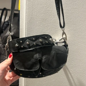 Svart axelväska med nitar från Unlimit - Säljer en cool svart axelväska från Unlimit. Väskan är i skinn och har snygga nitar som detaljer. Perfekt för en rockig look! Den har en dragkedja upptill och är i mycket bra skick. Passar perfekt för både vardag och fest.