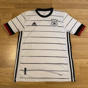 Vit Tyskland fotbollströja från 21/22 - Säljer en snygg Tyskland fotbollströja från Adidas med svarta ränder och detaljer. Tröjan har korta ärmar och en rund halsringning. Perfekt för fotbollsfans!