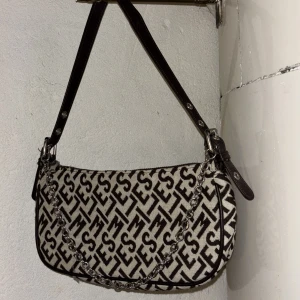 Mönstrad axelväska med kedjedetalj - Snygg axelväska med ett iögonfallande mönster i svart och beige. Väskan har en kedjedetalj framtill och justerbar axelrem i mörkt skinn. Perfekt för att ge din outfit en trendig touch.