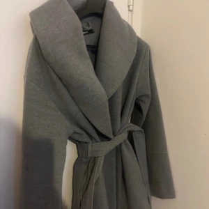 Grå kappa från BikBok - Snygg grå kappa från BikBok med ett brett skärp i midjan. Perfekt för kyligare dagar med sin stilrena design och långa ärmar. Ett måste i garderoben för en chic look.