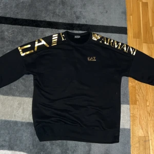 Svart tröja  EA7 Armani - Snygg svart tröja från EA7 Armani med guldtryck på axlarna och bröstet. Perfekt för en stilren look. Använd ett fåtal gånger och är helt felfri. Strl M 