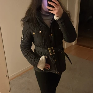 Svart quiltad jacka med bälte - Snygg svart quiltad jacka med bälte och guldiga knappar. Jackan har en figursydd passform och är perfekt för kyligare dagar. Den har långa ärmar och en stilren design med praktiska fickor. Är ifrån Chiara forthi, storlek M