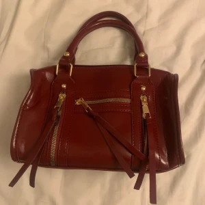 Röd handväska med dragkedja - Snygg röd handväska i skinn med guldfärgade detaljer och dragkedjor. Perfekt för att ge en färgklick till din outfit. Väskan har dubbla handtag och en praktisk ficka framtill.