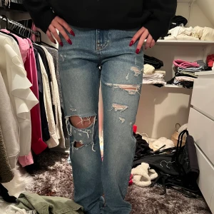 Blå slitna jeans - Säljer ett par blå jeans med slitningar och hål för en trendig look. De har en hög midja och en straight passform. Perfekta för en avslappnad stil.