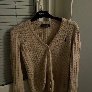 Beige kabelstickad kofta från Ralph Lauren - Säljer en stilren beige kabelstickad kofta från Ralph Lauren. Koftan har en v-ringad design med knappar framtill och det ikoniska logotypbroderiet på bröstet. Perfekt för en klassisk och elegant look. Orginal priset 1100 det är M men sitter som S