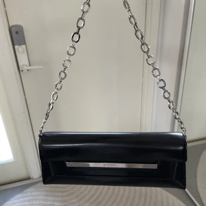 Svart axelväska från DKNY - Snygg svart axelväska från DKNY med en stilren design. Väskan har en kedjerem och en metallplatta med logotyp. Perfekt för att ge en elegant touch till din outfit.