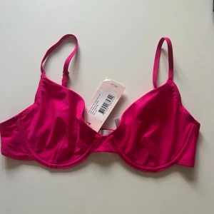 Neonrosa bikiniöverdel från Nelly - Snygg neonrosa bikiniöverdel från Nelly med bygel och justerbara axelband. Helt ny med prislapp kvar. 🩷 Storlek 75D.  Den har en elegant gulddetalj baktill som ger en extra touch av stil.