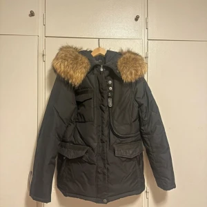 Svart dunjacka från Hollies - Snygg svart dunjacka från Hollies med avtagbar fuskpäls på huvan. Jackan har flera praktiska fickor och stängs med dragkedja och knappar. Perfekt för kalla vinterdagar. Är i mycket bra skick.