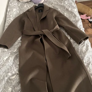 Brun kappa från MNG - Elegant brun kappa från MNG med knytbälte i midjan. Perfekt för kyligare dagar med sin långa design och klassiska snitt. Kappan har långa ärmar och en stilren krage.