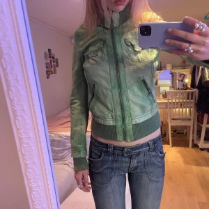 Grön skinnjacka med dragkedja - Snygg grön skinnjacka med dragkedja och ribbade muddar. Jackan har två bröstfickor med knappar och två sidofickor med dragkedja. Perfekt för en cool och avslappnad stil.