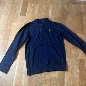 Mörkblå tröja från Lyle & Scott - Säljer en stilren mörkblå tröja från Lyle & Scott med dragkedja vid halsen och ett broderat gult emblem på bröstet. Perfekt för en avslappnad och snygg look. OBS de är till 12-13 åringar 