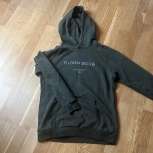 Grå hoodie från Björn Borg - Snygg mörk grön hoodie från Björn Borg med tryck 'Stockholm 1956' på bröstet. Perfekt för en avslappnad stil med en bekväm passform och huva. Storleken är 158-164