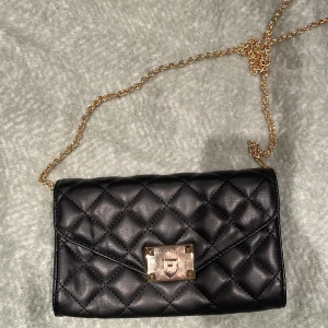 Svart quiltad axelväska med guldkedja - Snygg svart quiltad axelväska med guldkedja och ett elegant lås framtill. Väskan har en praktisk dragkedja inuti för extra säkerhet. Perfekt för att ge din outfit en stilfull touch.