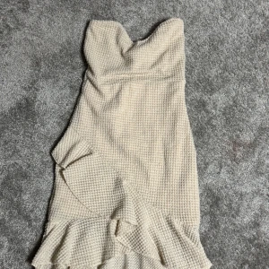 Beige axelbandslös klänning - Snygg beige axelbandslös klänning med volangdetaljer. Perfekt för en stilren look med en twist. Materialet har en texturerad yta som ger en unik känsla.