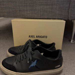 Mörk blåa sneakers från Axel Arigato - Snygga blåa sneakers från Axel Arigato med en stilren design. Skorna har en blå broderad fågel på sidan och vit sula. Perfekta för en trendig look. Får även med orginalkartongen! 