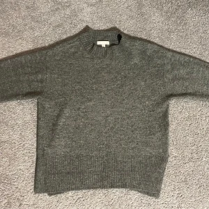 Grå stickad tröja från H&M - Mysig grå stickad tröja från H&M i storlek XS. Tröjan har en klassisk design med ribbstickade muddar vid ärmslut och nederkant. Perfekt för kyligare dagar.