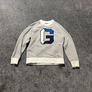 Stickad tröja från Gant L - Säljer en helt oanvänd stickad grå tröja från GANT med ett stort blått och svart G på framsidan. Tröjan har långa ärmar och ribbade muddar vid ärmslut och nederkant. Storlek L men passar även för M. Nypris ligger runt 800-1000kr.