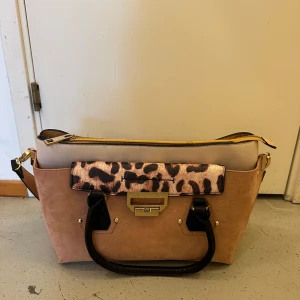 Beige axelväska med leoparddetaljer - Snygg beige axelväska med leopardmönstrad detalj och svarta handtag. Väskan har en dragkedja upptill och en justerbar axelrem. Perfekt för att ge en trendig touch till din outfit.