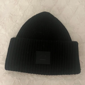 Svart ribbad mössa från Acne Studios - Snygg svart ribbad mössa från Acne Studios med ett stilrent märke framtill. Perfekt för att hålla värmen under kyliga dagar.