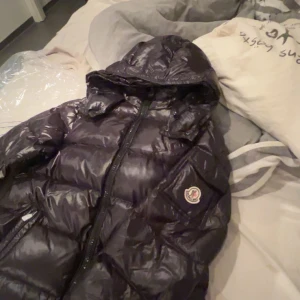 Svart dunjacka från Moncler - Säljer en snygg svart dunjacka från Moncler med huva och dragkedja. Jackan har ett glansigt yttre och Moncler-logga på ärmen. Perfekt för kyliga dagar.