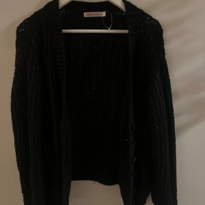 Svart stickad kofta från Indiska - Mysig svart stickad kofta från Indiska. Perfekt för kyliga dagar med sin öppna design och långa ärmar. Koftan har en fin textur och är lätt att matcha med olika outfits.