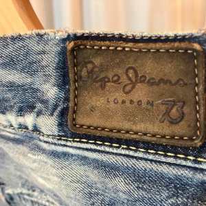 Blå slitna jeans från Pepe Jeans - Säljer ett par coola blå jeans från Pepe Jeans med slitna detaljer och en avslappnad look. Perfekta för en trendig och avslappnad stil. De har en klassisk femficksdesign och knappgylf. 31 midja 32 längd.