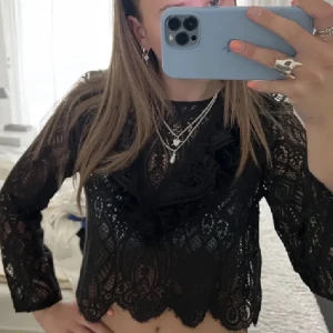 Svart spetsblus - Snygg svart spetsblus med långa ärmar och volangdetaljer. Perfekt för en elegant look. Blusen är genomskinlig och har en croppad passform.