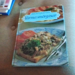 Kokbok: Varma smörgåsar - En kokbok med fokus på varma smörgåsar. Omslaget visar en aptitretande bild av en smörgås med svamp och persilja, serverad på en blåvit tallrik. Perfekt för den som vill utforska olika recept på varma mackor.