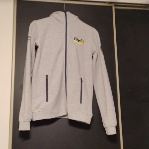 Grå hoodie med HV71-logga - Säljer en grå hoodie med dragkedja och HV71-logga i blått och gult. Tröjan har långa ärmar och två fickor med dragkedjor framtill. Perfekt för fans av HV71! 🏒 Använd en gång. Pris kan diskuteras