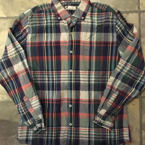 Rutig skjorta från Dressmann  - Säljer en klassisk rutig skjorta från Dressmann i storlek L. Skjortan har ett färgglatt mönster i blått, rött och grönt med vita knappar.Har en ficka på vänster sida framtill. Tillverkad i 100% bomull, perfekt för en avslappnad stil.