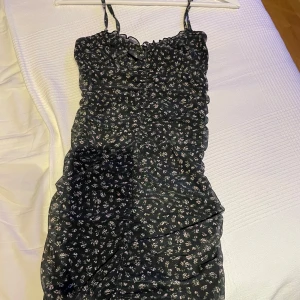 Svart blommig klänning med smala axelband - Använd 1 gång. Snygg svart klänning med små vita blommor och smala axelband från H&M. Klänningen har en åtsittande passform med rynkade detaljer framtill som ger en smickrande siluett. I storlek XS men har stretchigt material.